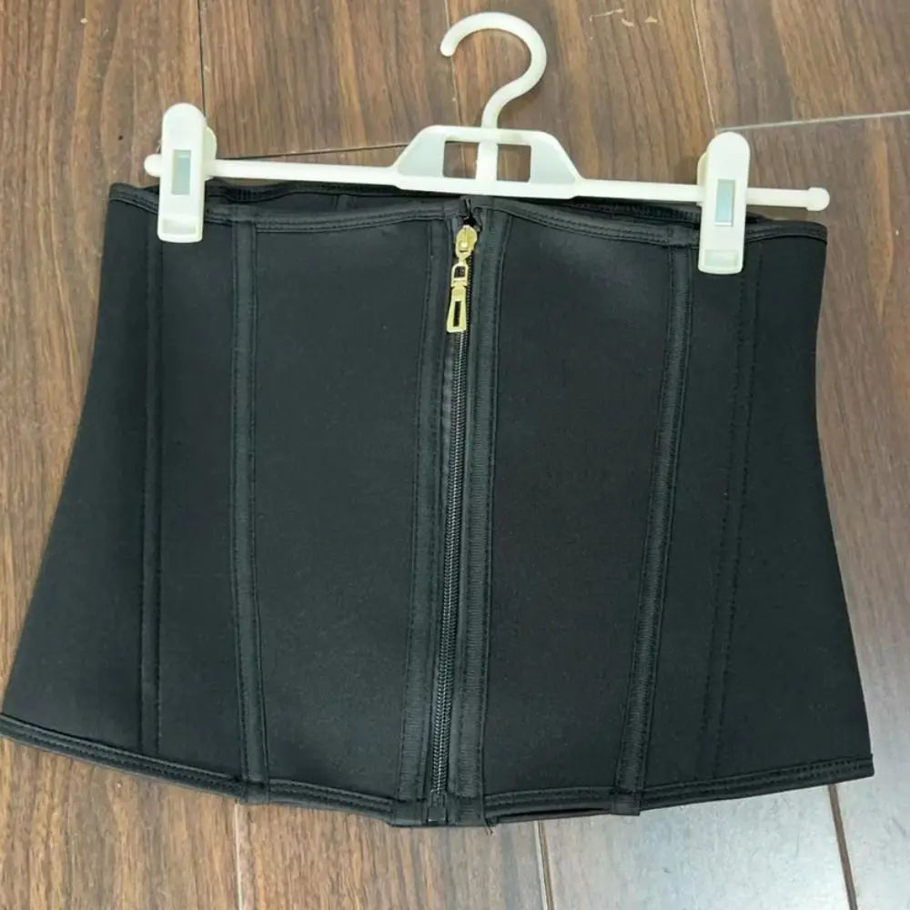 Black waist trainer with a gold zipper on a hanger against a wooden background
